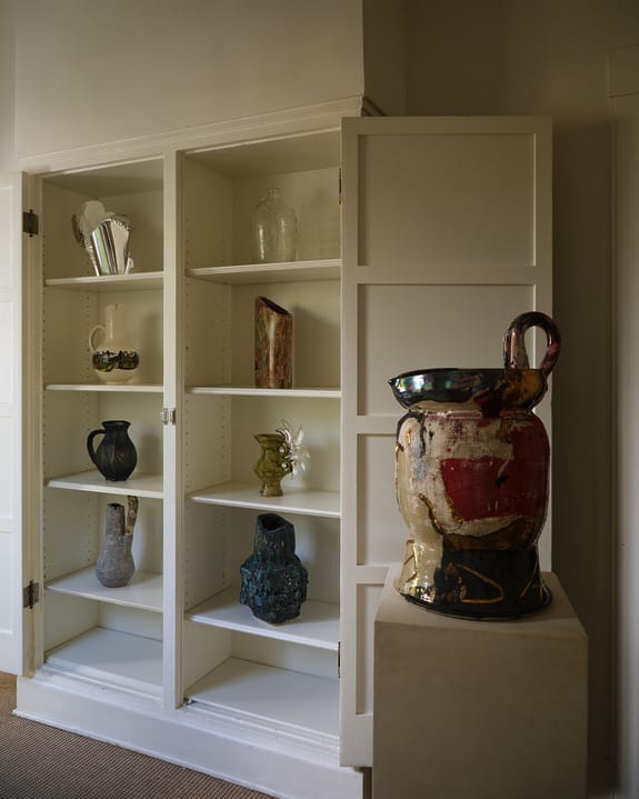 Open white cabinet displaying various ceramic vases and artworks with a large, colorful vase on a pedestal beside it.