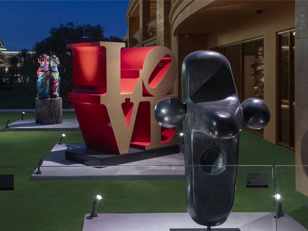 Outdoor art installation featuring a large red "LOVE" sculpture, abstract stone figures, and manicured green lawn at twilight.
