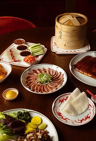 An assortment of Chinese dishes on a table featuring sliced duck, pancakes, condiments, and a bamboo steamer basket.