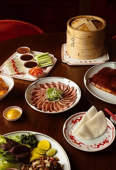 An assortment of Chinese dishes on a table featuring sliced duck, pancakes, condiments, and a bamboo steamer basket.