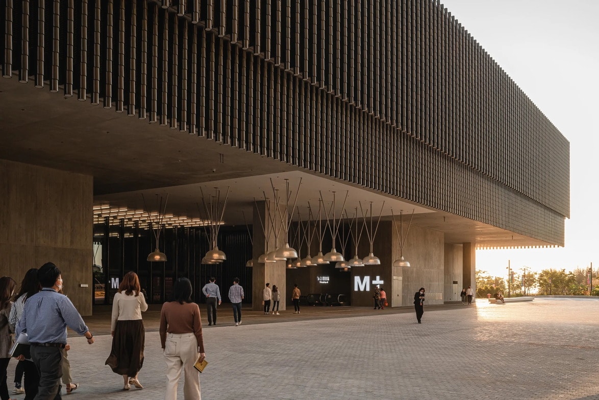 People walking towards a modern, architecturally striking building with a large overhang and suspended lamps.
