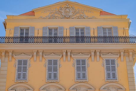 Ornate yellow building facade with decorative frieze, shuttered windows, and a black metal balcony against a clear blue sky.