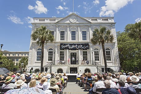 Historic building hosting outdoor event with crowd seated under clear blue sky.
