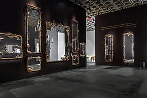 Art installation with ornate mirrors and dark walls in a gallery space with subtle lighting.