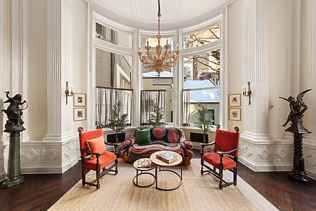 Luxurious sitting room with large bay window, chandelier, ornate decor, red chairs, and view of neighboring buildings.