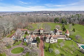 Aerial view of a large mansion with red roofs, surrounded by trees and open fields on a clear day.