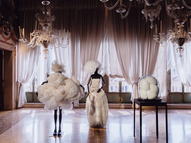 Elegant fashion display with avant-garde white dresses and chandeliers in a luxurious room with draped curtains.