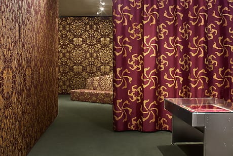 Ai Weiwei x Rubelli collaboration room at Milan Design Week with intricate patterned red curtains and wallpaper, featuring ornate golden designs, and a display case on the right.