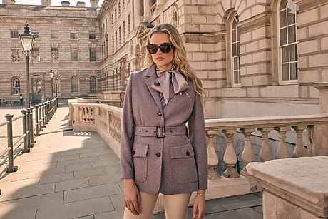 Woman in stylish blazer and sunglasses standing outdoors by a historic building.