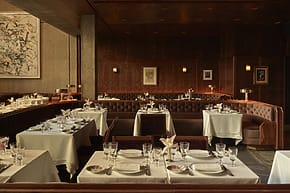 Elegant restaurant dining area with neatly set tables, white tablecloths, plush seating, and artwork on wood-paneled walls.