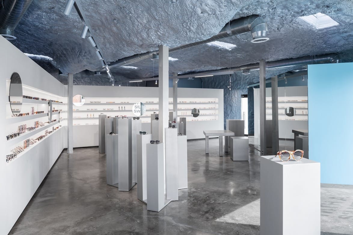 Modern eyewear store interior with sleek gray displays, glasses showcased on stands, and a textured ceiling design.
