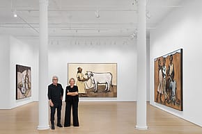 Two people standing in an art gallery with large paintings depicting figures and a cow on the white walls.