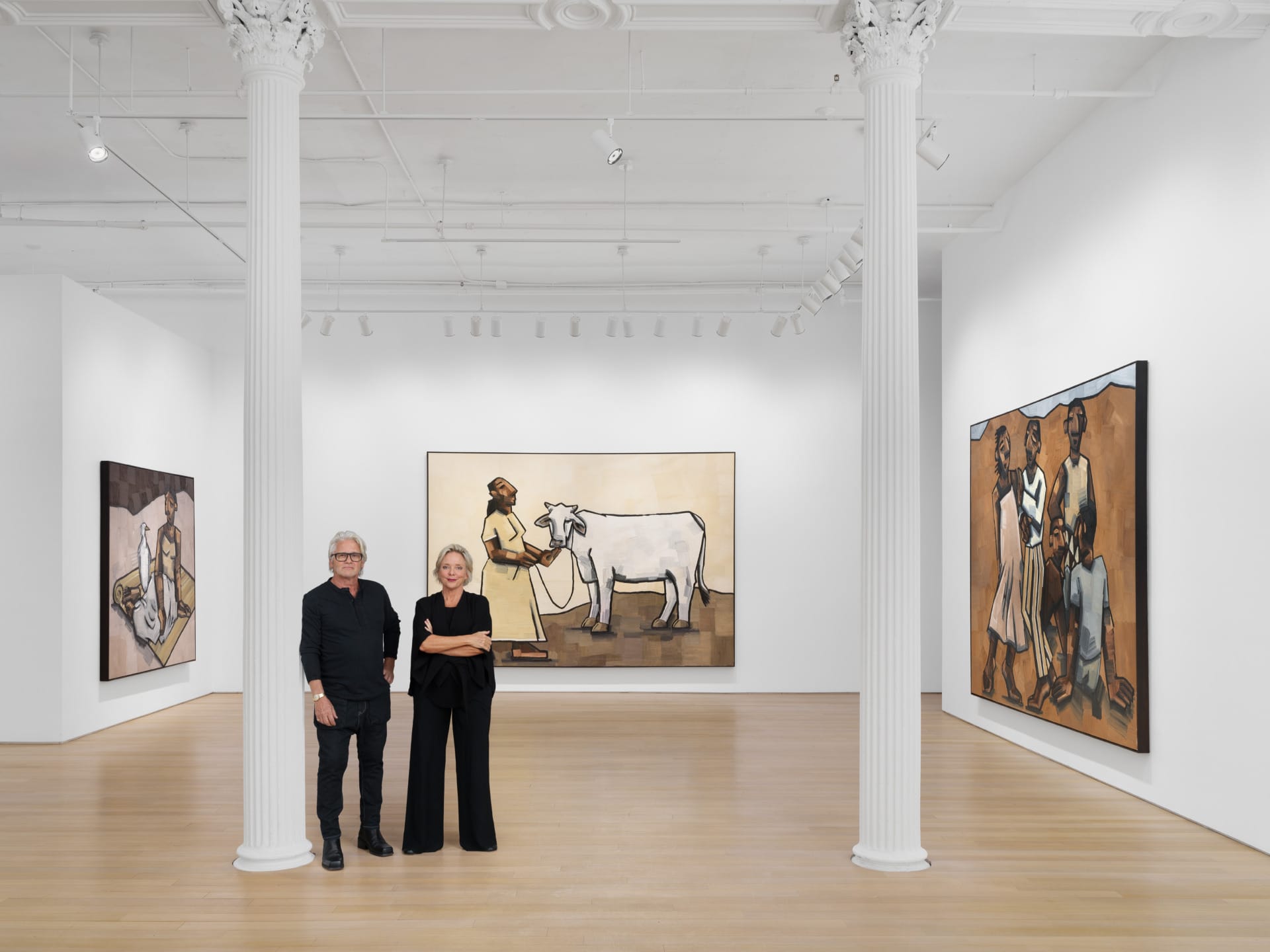 Two people standing in an art gallery with large paintings depicting figures and a cow on the white walls.