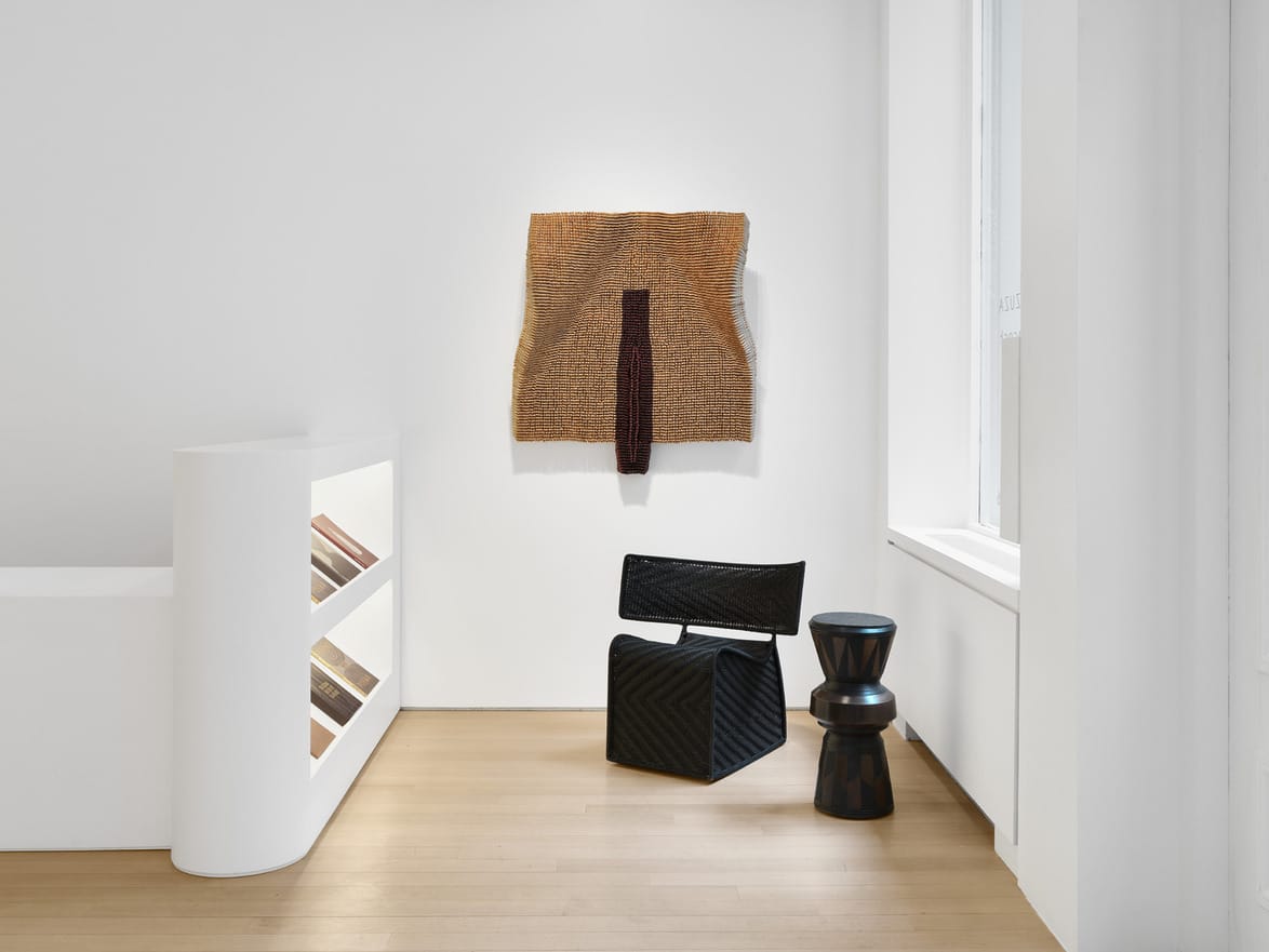 Modern art exhibit with woven textile on wall, black chair, and wooden stool in a bright, minimalist gallery space.