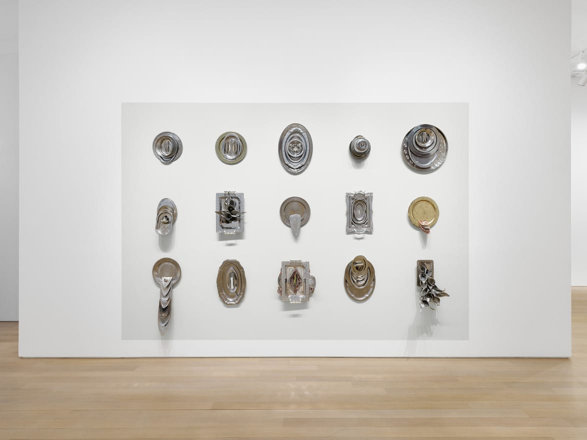 Wall-mounted abstract sculptures on a gallery wall displayed in a grid pattern with various shapes and textures.