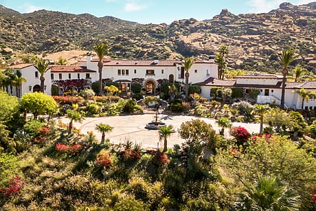 A luxurious Mediterranean-style mansion surrounded by lush gardens with a mountainous backdrop under a clear blue sky.