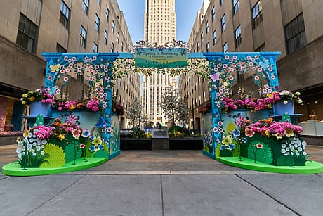 Colorful floral display with "Spring is Blooming" sign set in a bustling city plaza with tall buildings in the background.