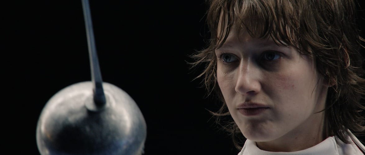 Person in fencing gear focusing intensely, with blurred fencing sword in foreground.