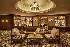 Elegant hotel lobby with plush seating, ornate ceiling, and built-in bookshelves warmly illuminated by ambient lighting.