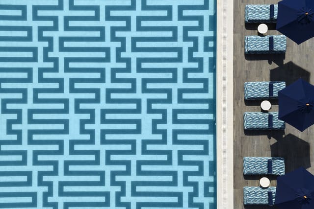 Aerial view of a patterned swimming pool beside lounge chairs and umbrellas on a sunny day.