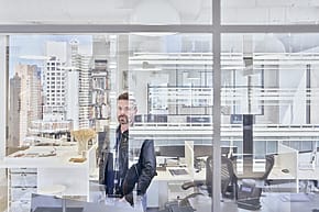 Man in a modern office standing by a window, with urban buildings visible in the background.