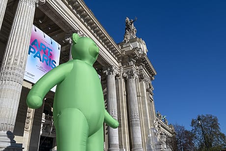 Giant green inflatable bear in front of a historic building with tall columns under a clear blue sky.