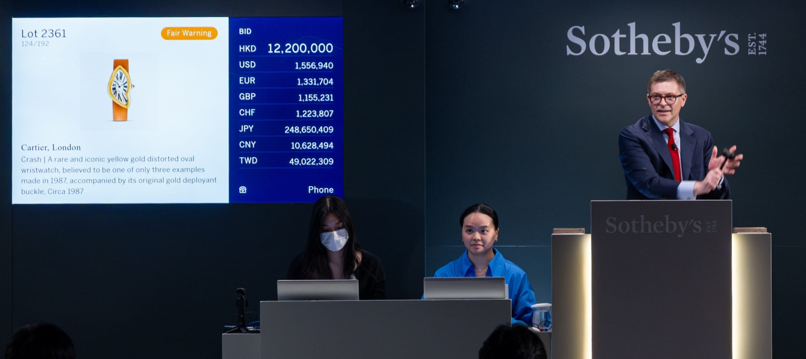 Auctioneer at Sotheby's with two assistants, leading a watch auction with bid details displayed on a screen.