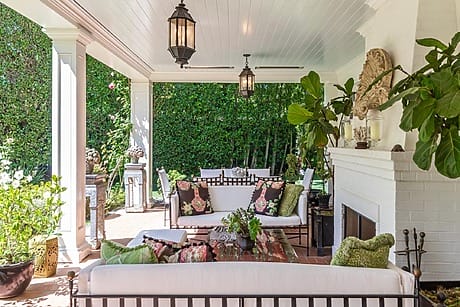 Covered outdoor patio with white sofas, colorful cushions, fireplace, hanging lanterns, and lush greenery in the background.
