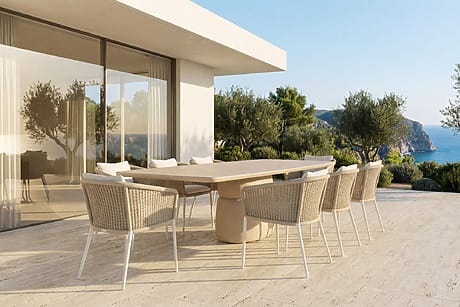 Modern patio with wicker chairs and large table, ocean view and greenery in the background, under a clear blue sky.