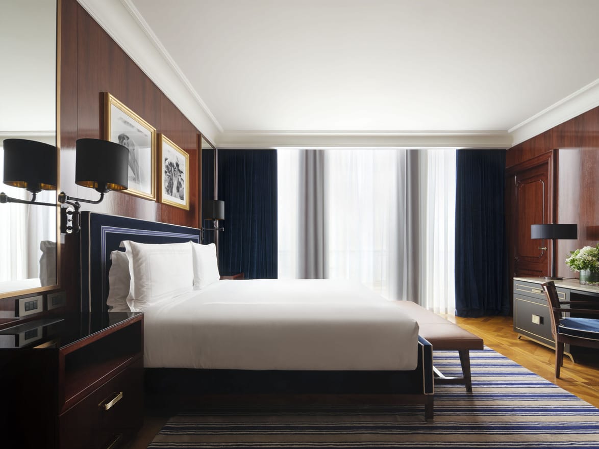 Elegant hotel room at Casa Cipriani Milan with a large bed, wooden furniture, striped rug, and a window with sheer curtains letting in natural light.
