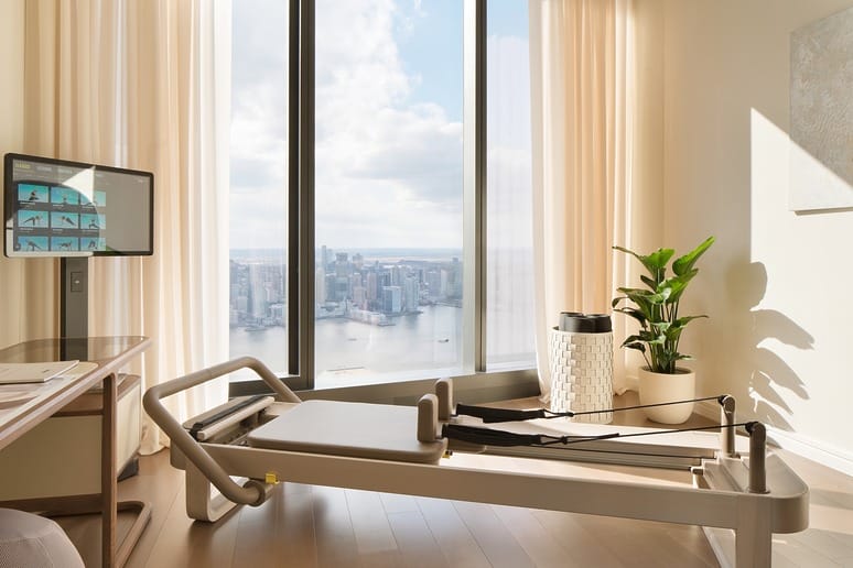 Modern exercise room with reformer, large windows, city skyline view, potted plant, desk, and a wall-mounted screen.