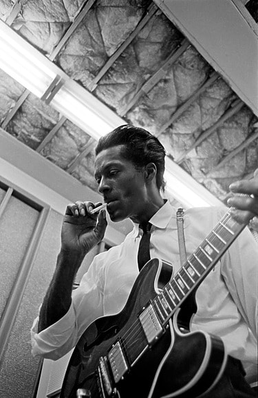 Musician Chuck Berry playing electric guitar in a recording studio, wearing a tie and white shirt, at a club, photographed by Don Bronstein.