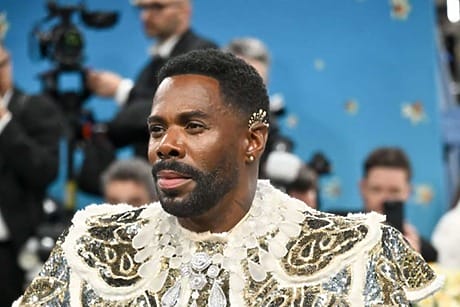 Person in ornate white and gold outfit with floral background, surrounded by people and cameras at an event.