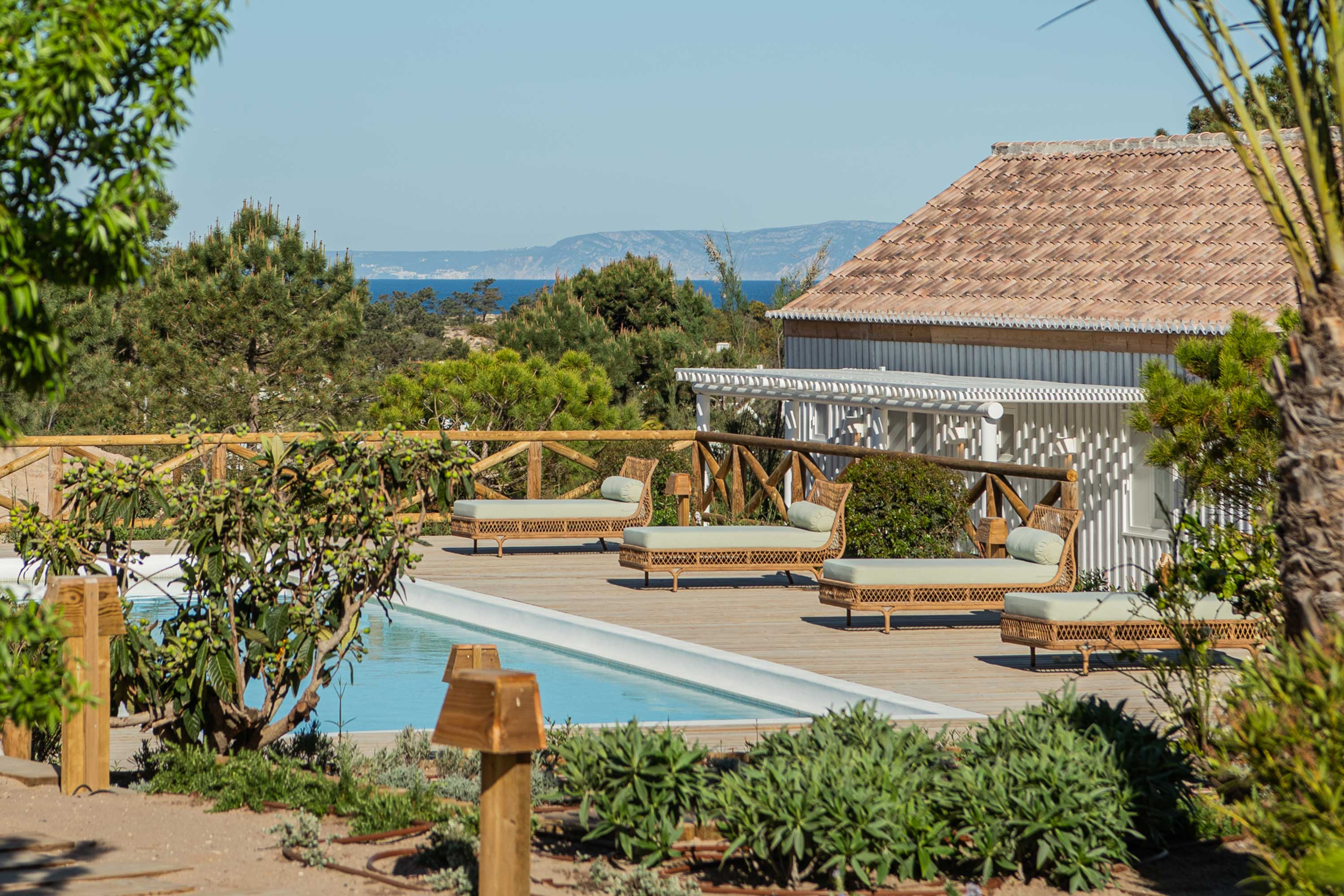 Scenic view of a luxury poolside area with lounge chairs, surrounded by greenery, and partial ocean view in the background.
