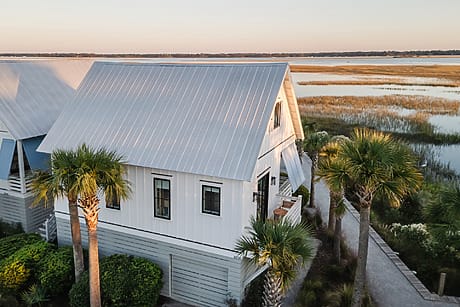 White coastal cottage with metal roof and palm trees near water and marshland, under a clear blue sky.