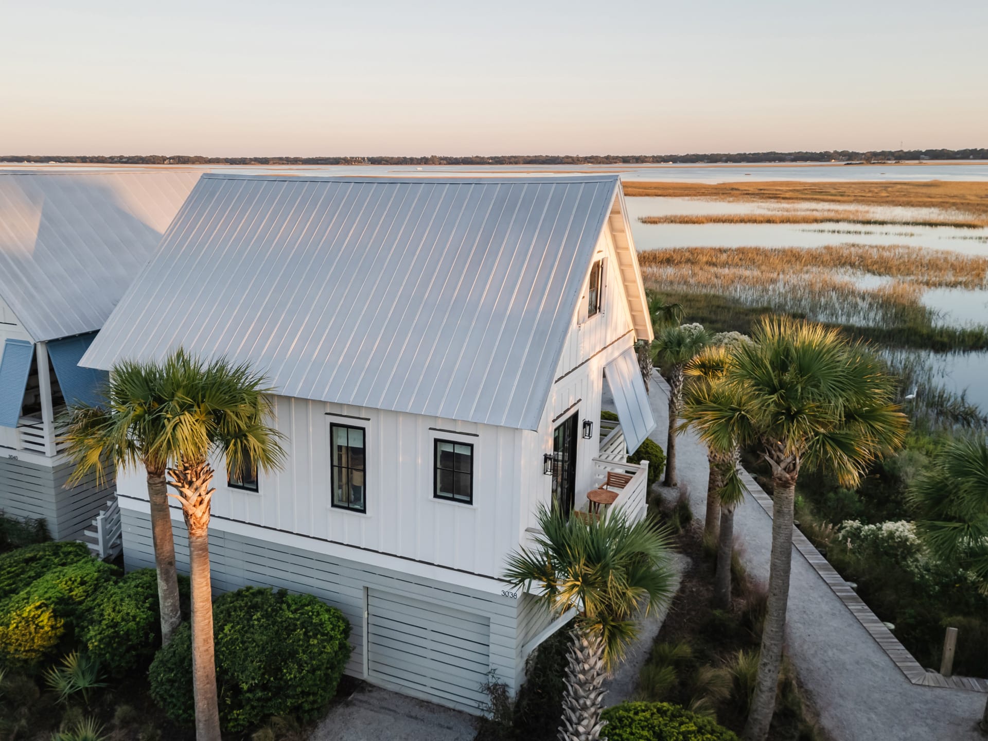 White coastal cottage with metal roof and palm trees near water and marshland, under a clear blue sky.