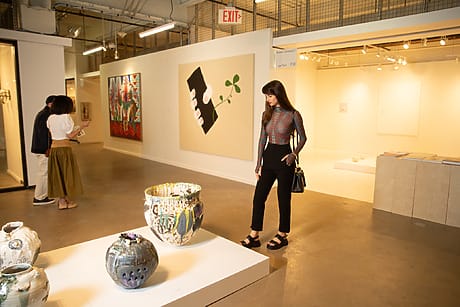 A woman in an art gallery observing pottery and wall art, with two people in the background.