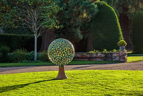Colorful spherical sculpture on a green lawn with lush trees and manicured garden in the background.