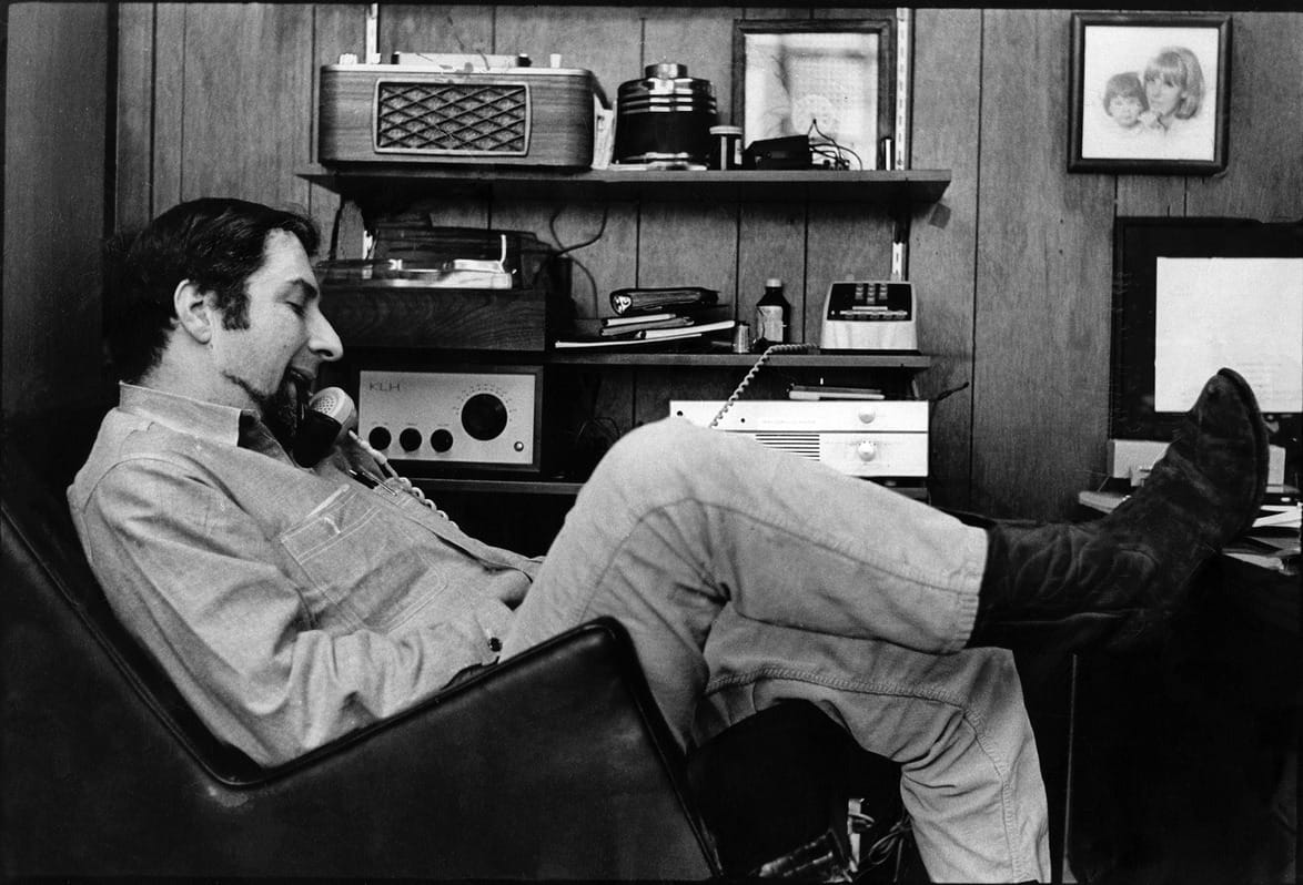Photographer Don Bronstein sitting in a chair, wearing boots, talking on a phone in a vintage office setting with radios and photos.