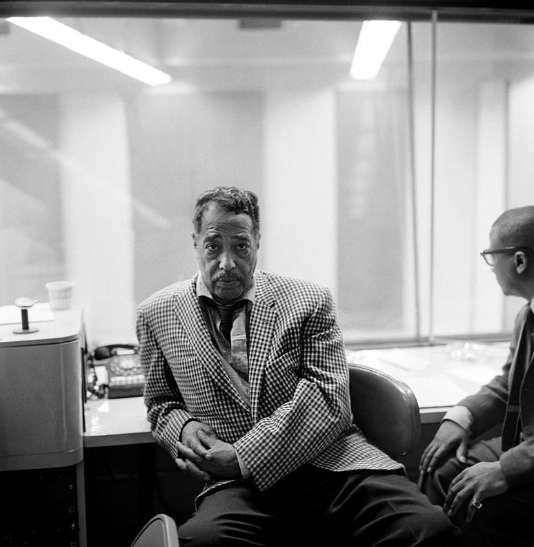 Duke Ellington sitting in a recording studio, with one leaning back in a chair wearing a checkered suit, looking at the camera, in a photo taken by Don Bronstein