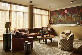 Cozy living room with a brown sectional sofa, patterned chairs, wooden table, and colorful wall tapestry.