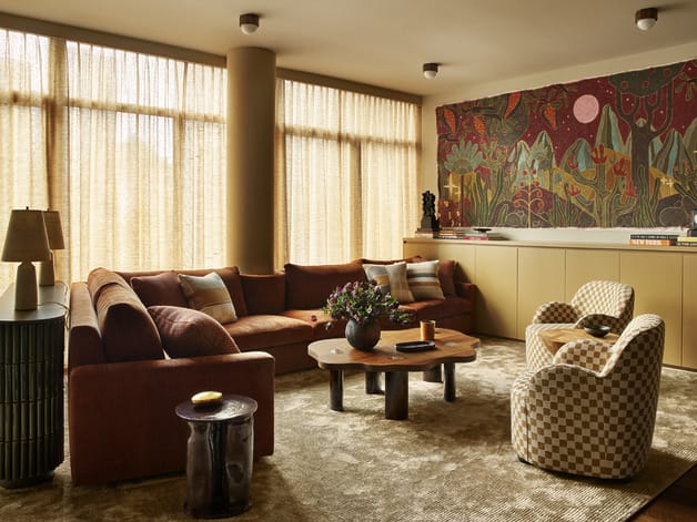 Cozy living room with a brown sectional sofa, patterned chairs, wooden table, and colorful wall tapestry.