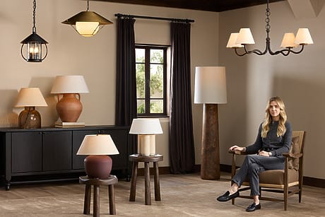 Woman sitting in living room with various stylish lamps, dark curtains, wood flooring, and ambient lighting.