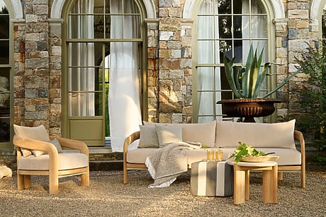 Outdoor patio set with beige cushions, wooden table, and armchair against stone wall and large potted plant.