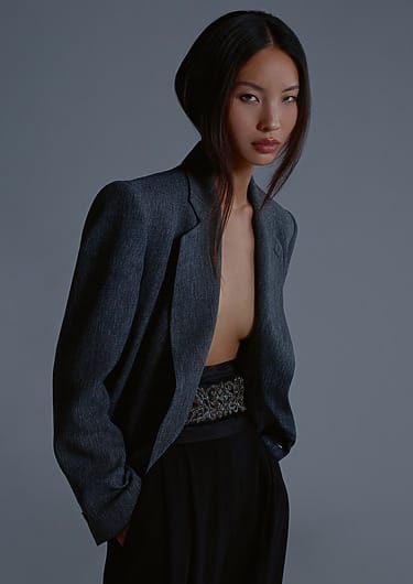 Woman in a partially open blazer with long hair, standing against a gray background, looking confidently at the camera.