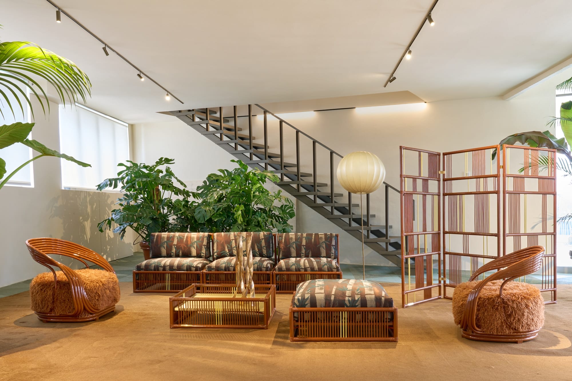 Modern living room with patterned sofa, wooden chairs, glass table, large plants, and staircase in a well-lit space.