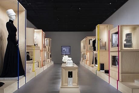Exhibition room with mannequins in elegant dresses and display boxes along both walls, showcasing various fashion items.