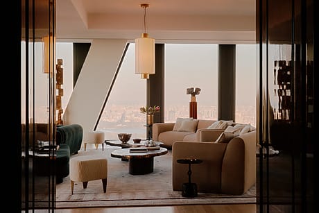 Modern living room with large windows, beige sofas, elegant lighting, and city skyline view.