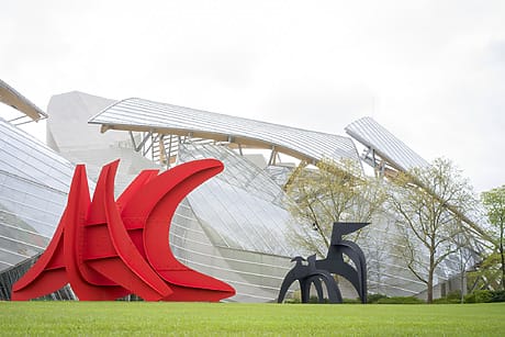 Red and black abstract sculptures on green grass with modern glass building and trees in the background.