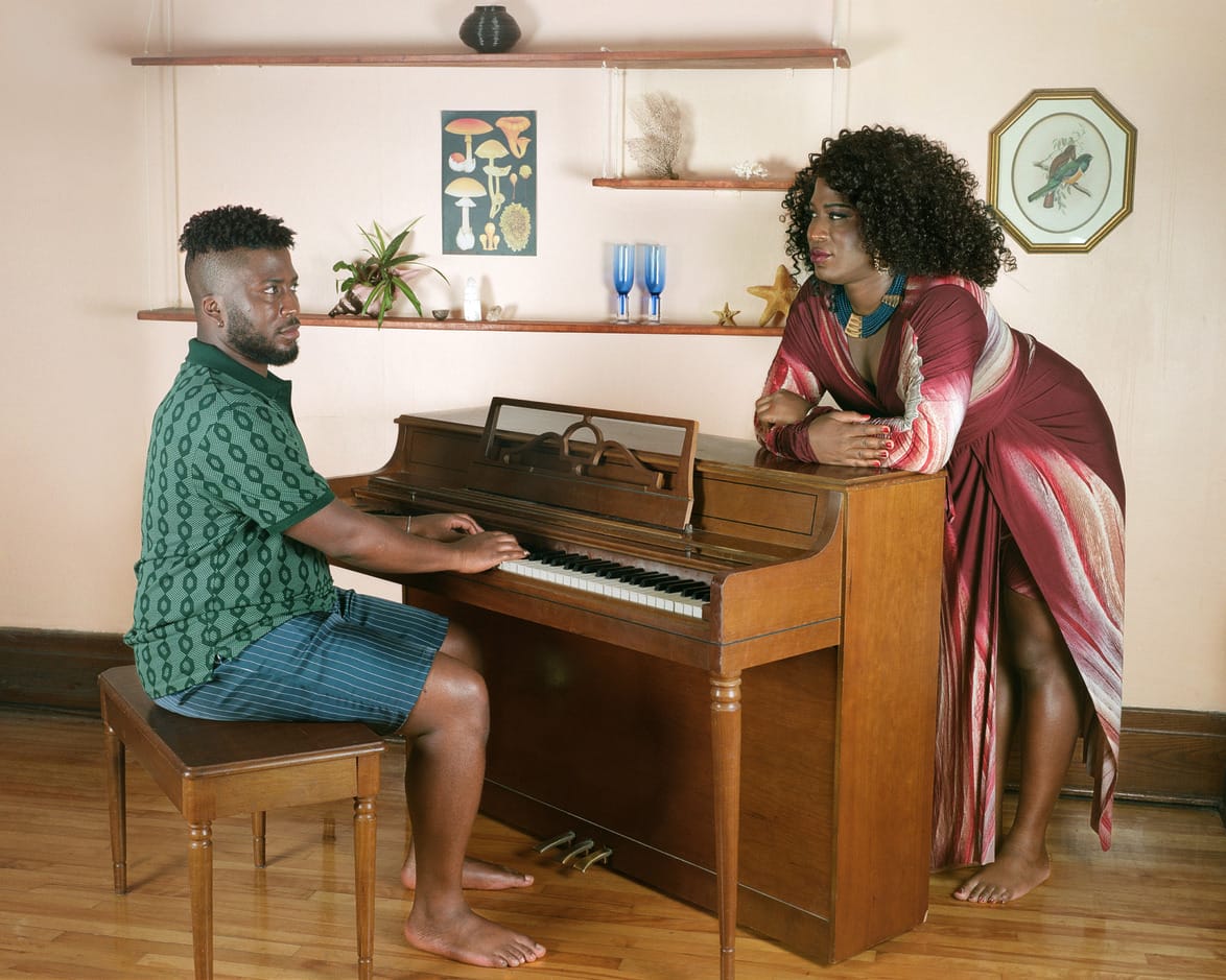 Same model portraying man playing piano while woman listens, standing against it in a cozy room with shelves and framed art, image taken by JJ Levine and displayed at AIPAD at the Park Avenue Armory in New York.